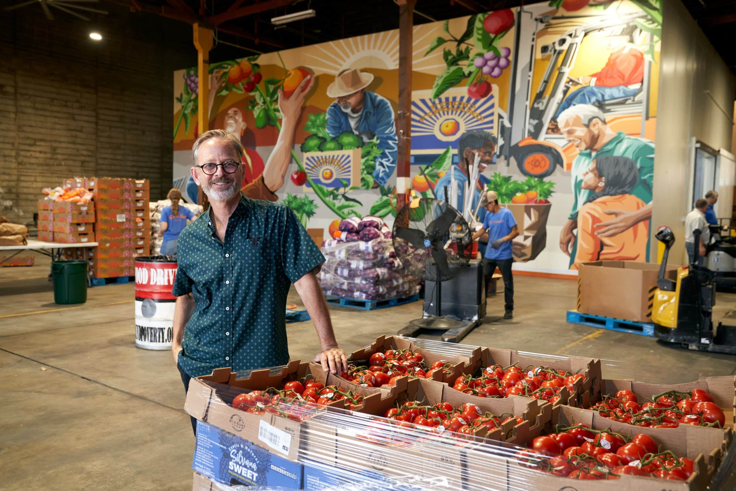A smiling man with glasses stands inside a food warehouse, resting his hand on boxes filled with red tomatoes. Behind him, volunteers sort produce, and a large colorful mural shows farmers, food, and community scenes on the wall.